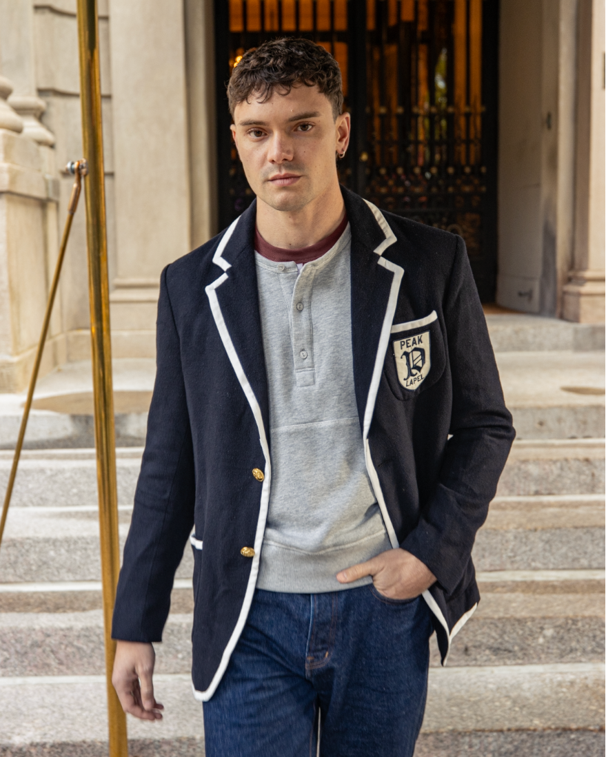 Man wearing a navy blazer with a school emblem, standing on steps in an urban setting.